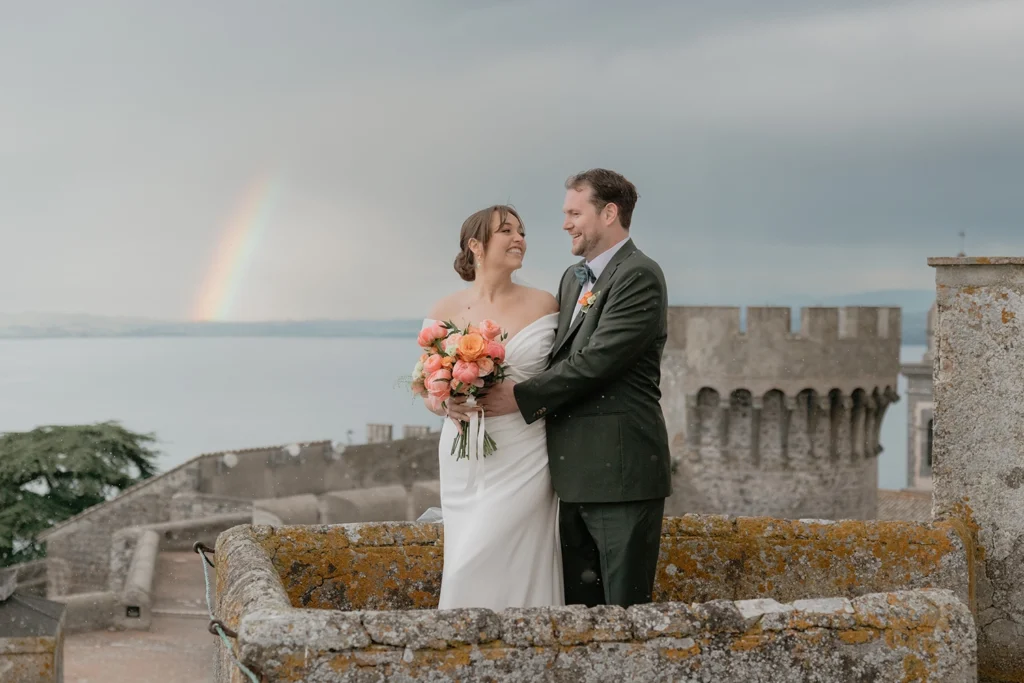 a couple is looking at each other at Castello Orsini Odescalchi di Bracciano and a rainbow is in the sky
