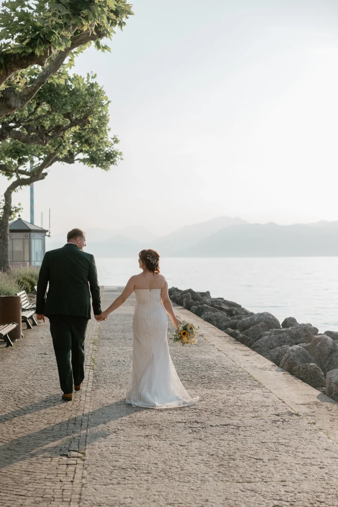 a newlywed couple is walking hand to hand on lake Garda shores